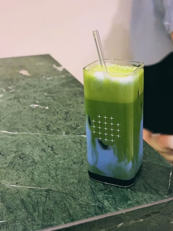 A layered matcha drink — vivid green over pale butterfly-pea blue — in a clear ONO star-grid cup on green marble