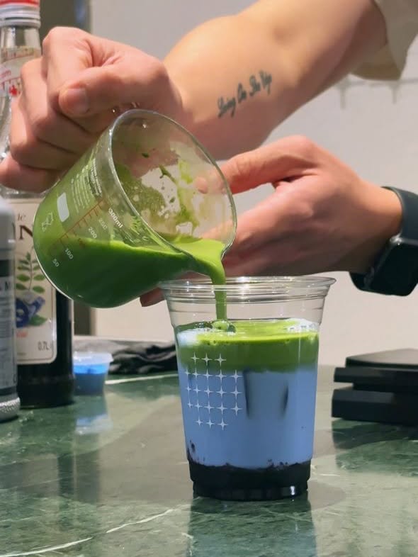 A barista pours vivid matcha over a butterfly-pea layer in a clear ONO star-grid cup on a green marble counter